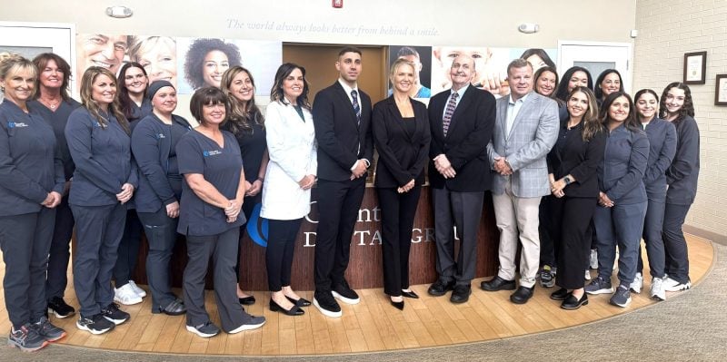 Large group photo of dental office team members wearing navy blue scrubs and professional business attire, standing together in modern office space with wood flooring