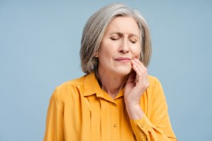 Middle-aged woman in yellow top holding her jaw in discomfort, representing dental pain or temporomandibular joint disorder